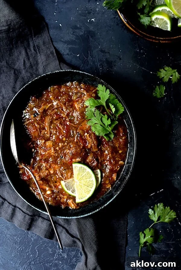Slow Cooker Colorado Green Chili in black bowl garnished with lime wedges and cilantro sprigs.