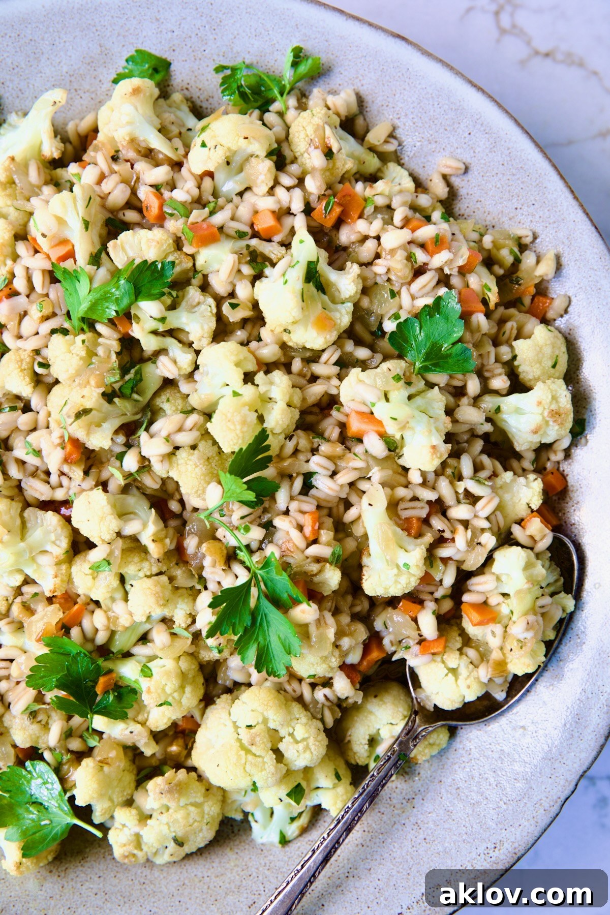 Close-up of a serving bowl filled with Barley Pilaf and Roasted Cauliflower, elegantly garnished with fresh parsley and a serving spoon.