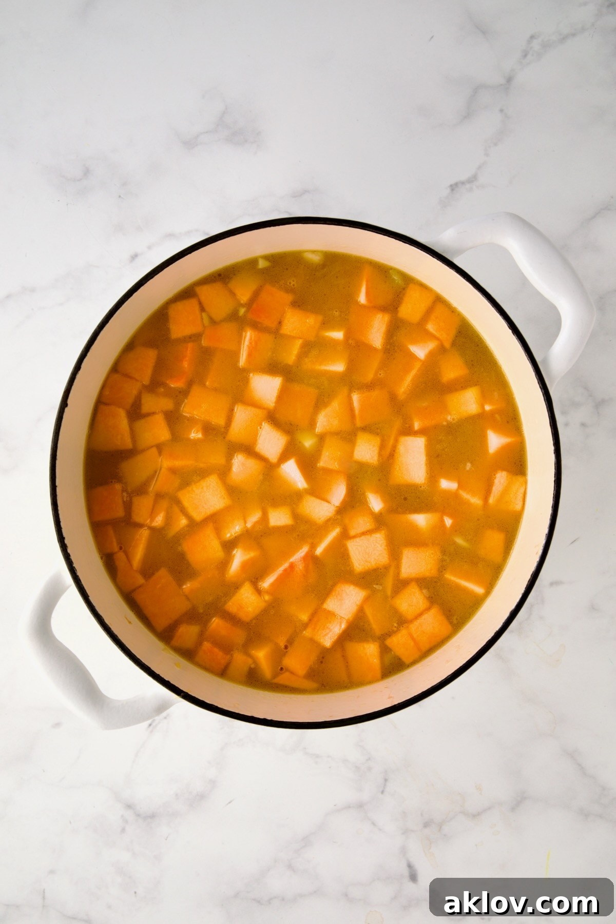 Cubed butternut squash and savory chicken broth carefully added to the sautéed vegetables in the white enamel Dutch oven, preparing for simmering.
