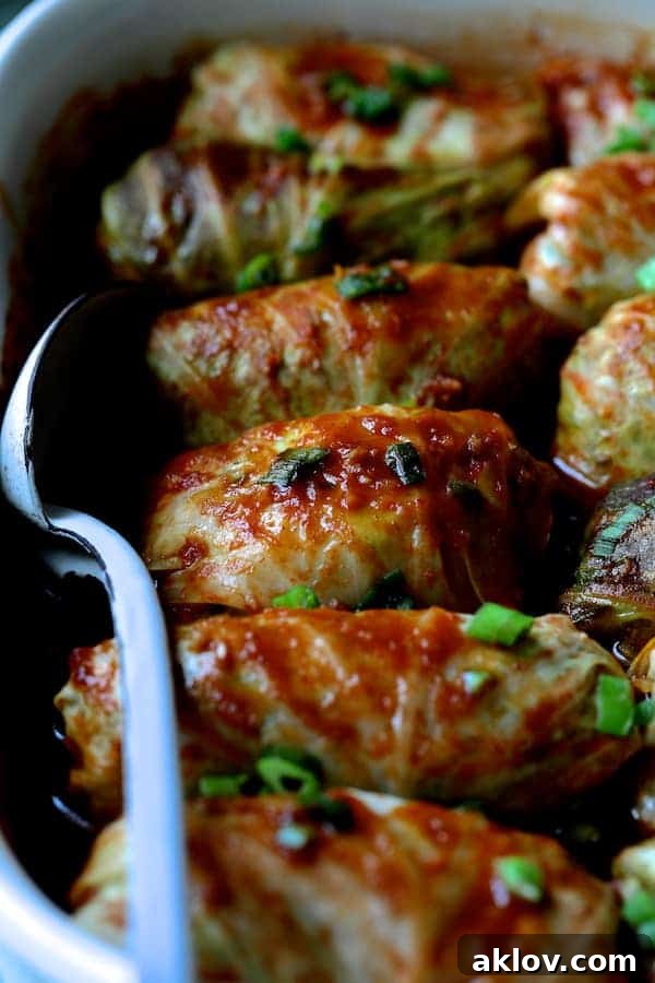 Asian Spiced Pork Cabbage Bundles 8 Close-up photo of cooked Spicy Asian Pork Cabbage Rolls glistening with sauce in a white baking dish, ready to be served with a white metal spoon.