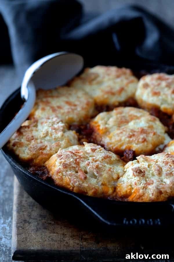 Close-up photo of Skillet Turkey Pinto Bean Chili with Pepper Jack Biscuit Topping in cast iron skillet with gray dish towel on wooden trivet. The baked, golden-brown biscuits sit atop the bubbling chili.