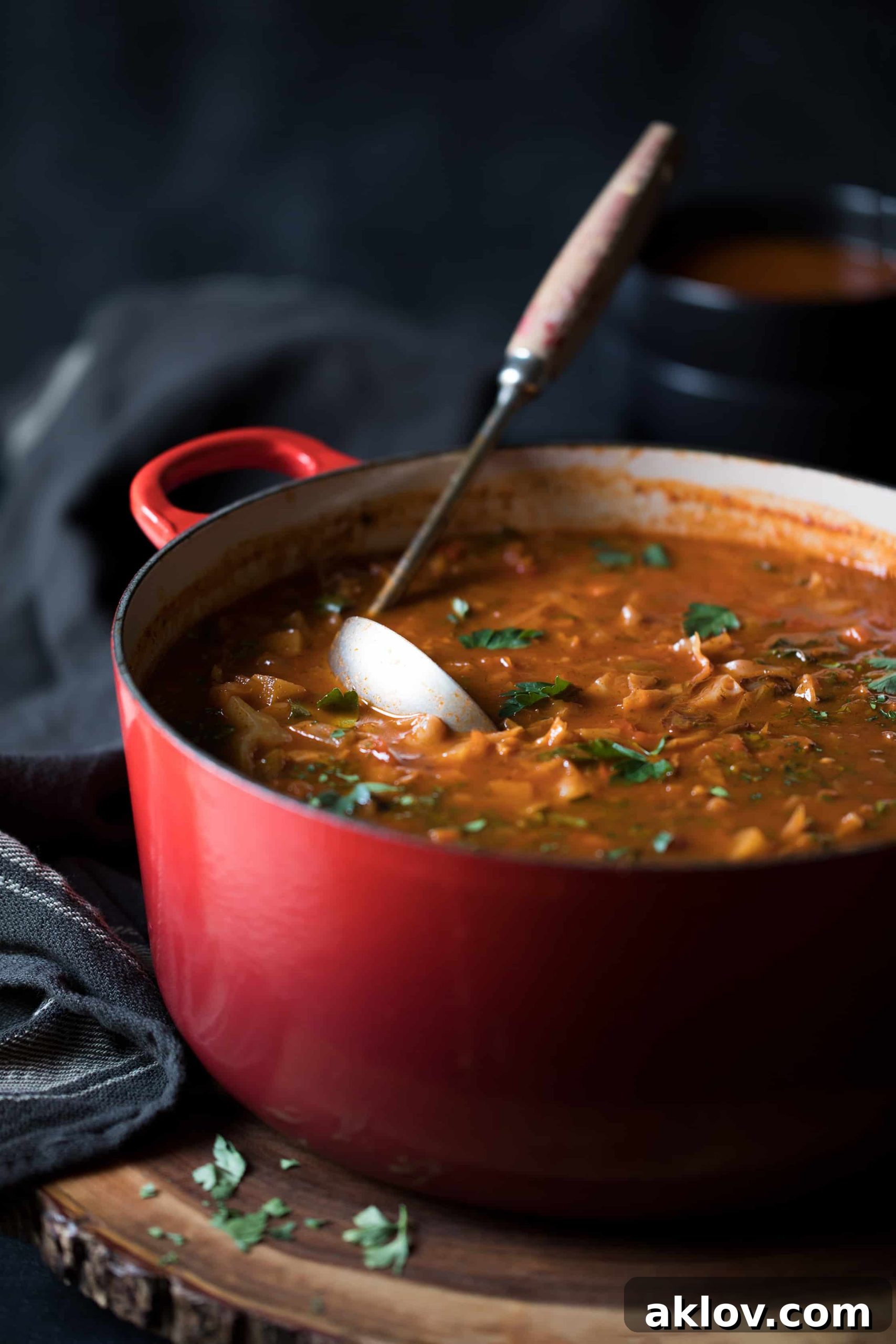 Rustic Cabbage and Root Vegetable Soup simmering in a red pot with a ladle, placed on a wooden trivet next to a striped towel.