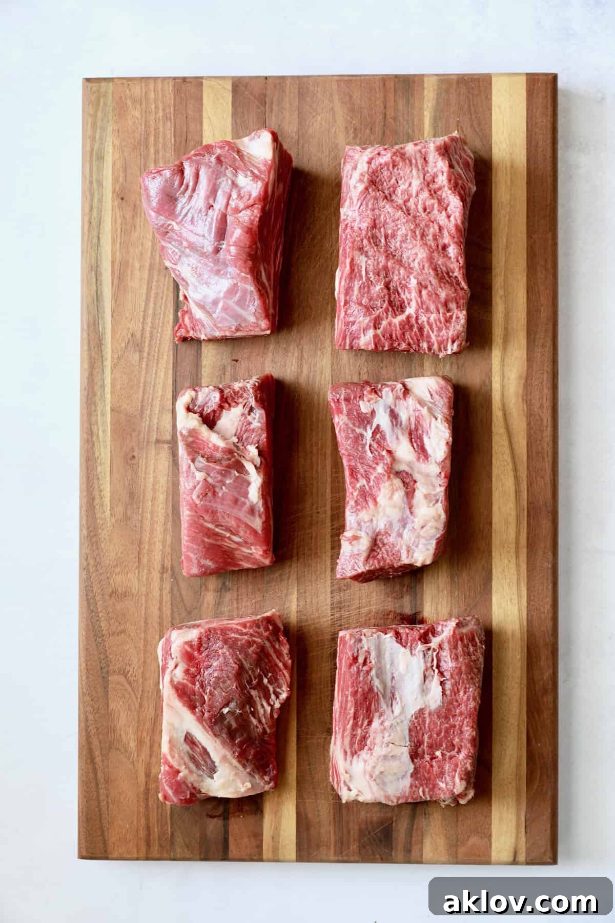Six uncooked, generously marbled beef short ribs are neatly arranged on a rustic wooden cutting board, ready for seasoning and searing.