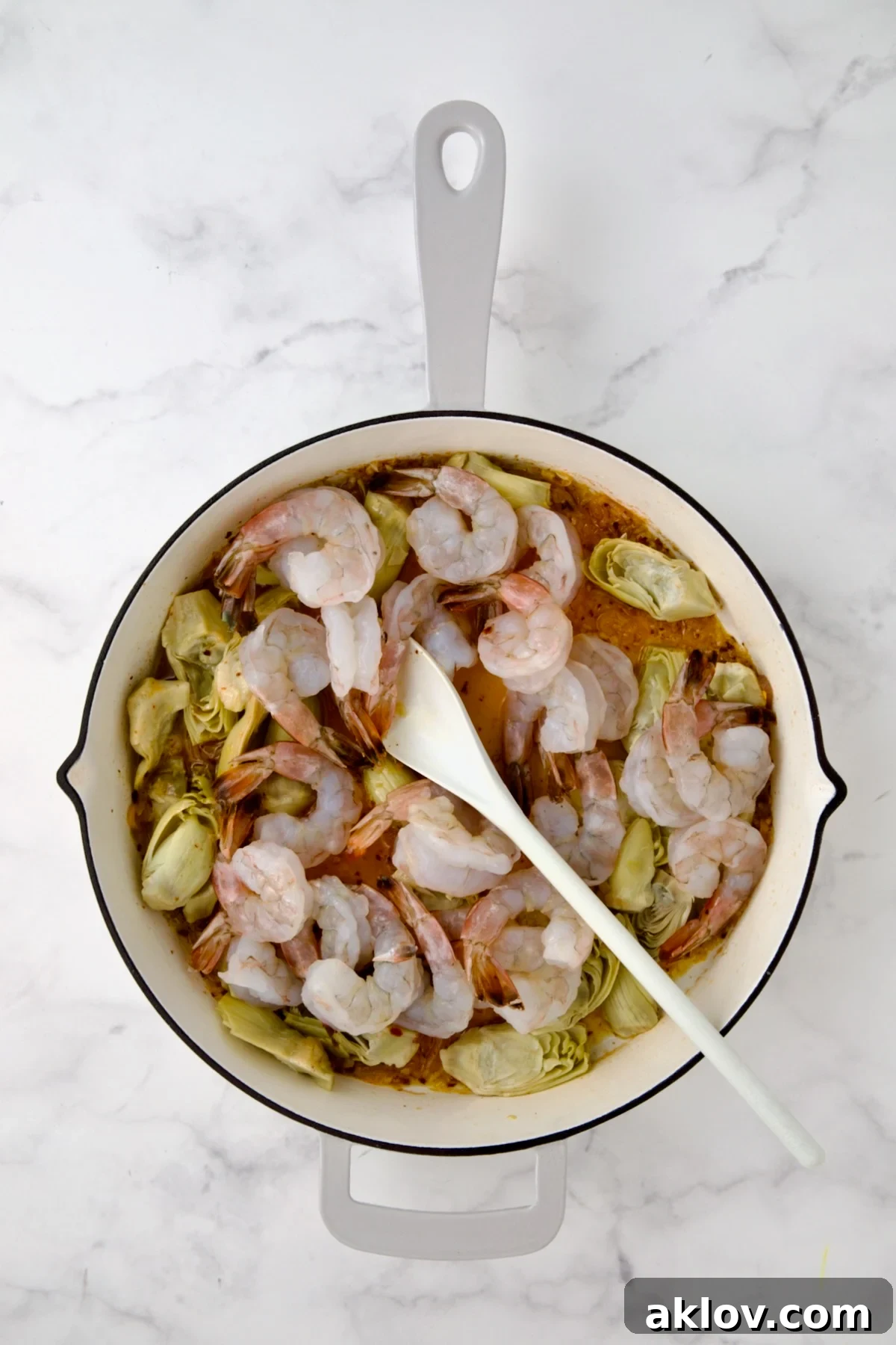 Shrimp added to artichokes in white enamel cast iron pan being stirred with white mixing spoon.