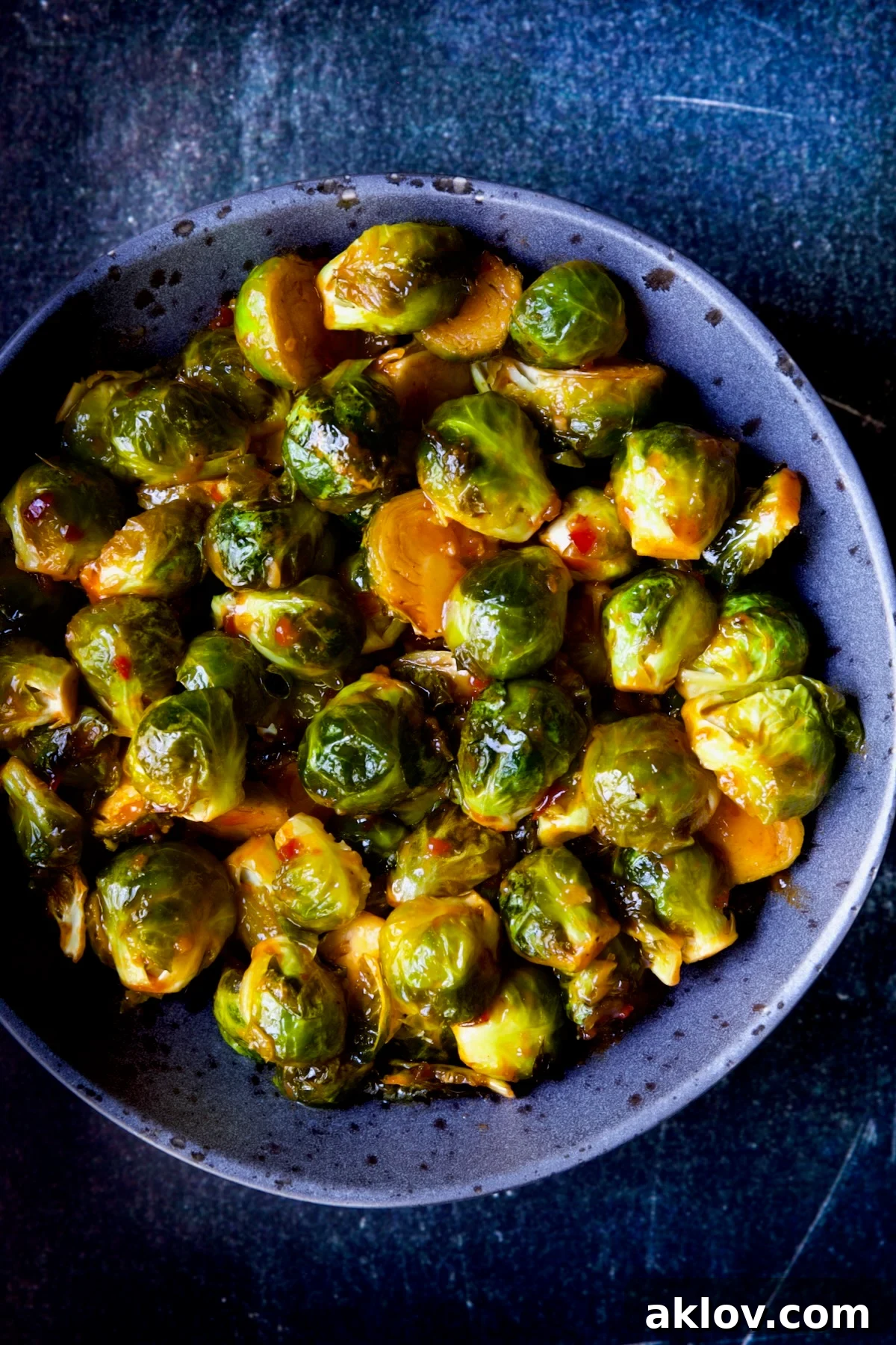 Glazed Sriracha Sweet Chili Brussels Sprouts 13 A close-up shot of Sriracha Sweet Chili Roasted Brussels Sprouts served in a gray speckled bowl, showing their glossy, caramelized texture.