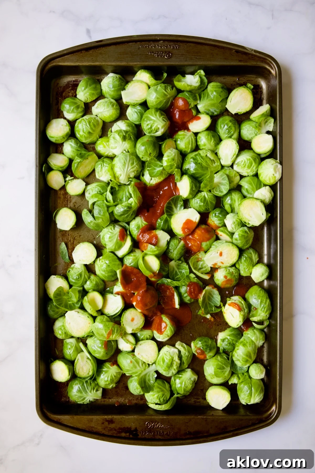 Glazed Sriracha Sweet Chili Brussels Sprouts 7 Halved Brussels sprouts on a baking sheet with Sriracha hot sauce being poured over them.