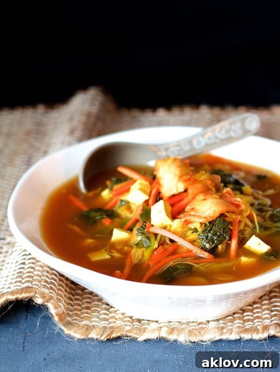 Photo of Asian Vegetable Soup with Tofu, Kimchi and Rice Noodles in white bowl on brown burlap and blue background, garnished with fresh herbs.