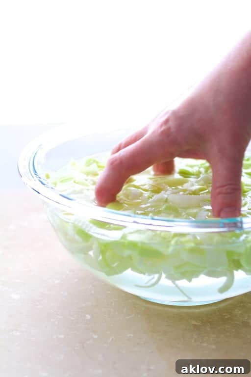 A hand gently swirling sliced leeks in a clear glass bowl of water to wash away dirt.