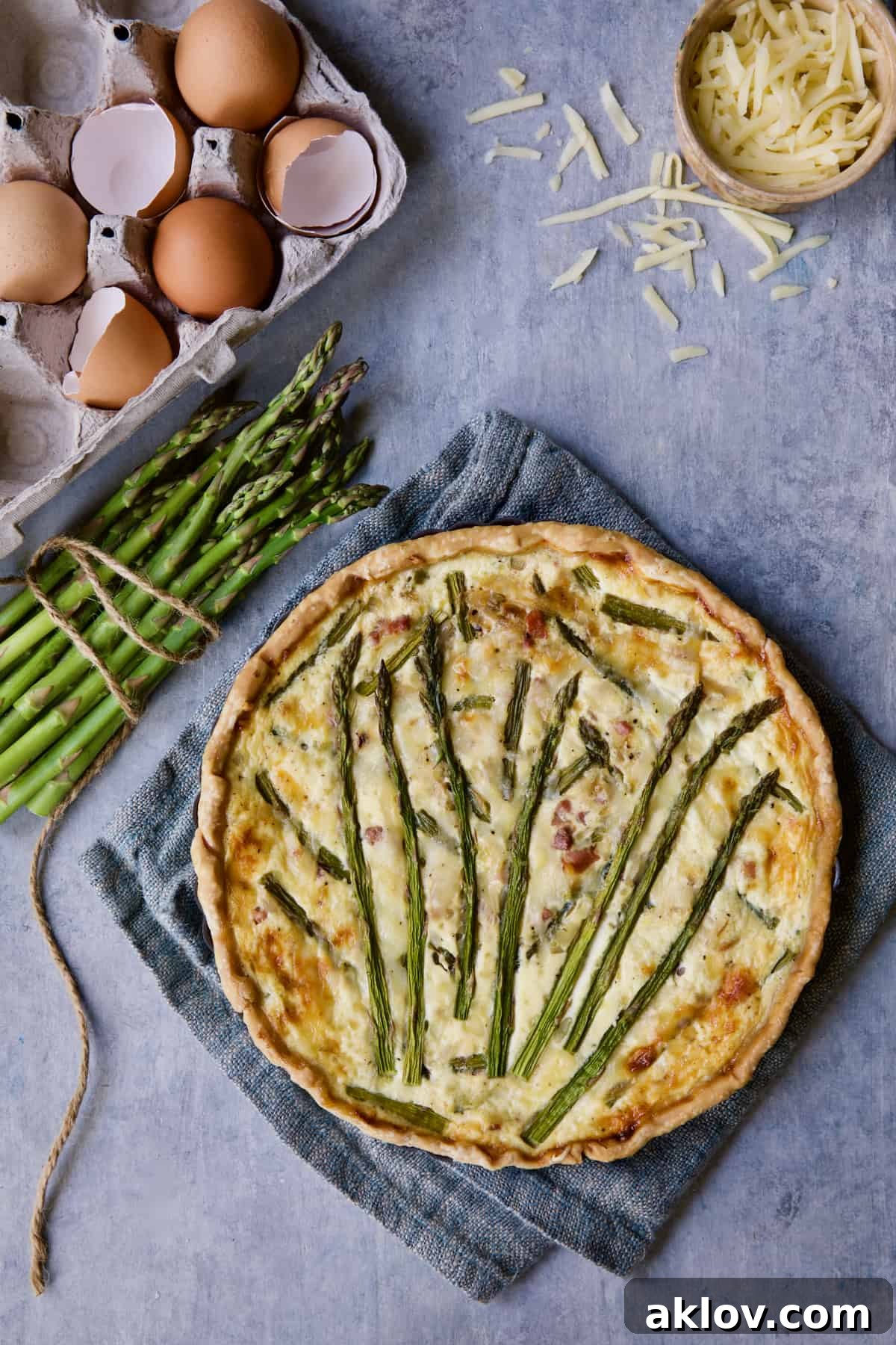 Savory Asparagus Tart with Prosciutto and Fontina 11 Photo of a freshly baked Asparagus Prosciutto and Fontina Tart, garnished with fresh asparagus, cheese, and a few scattered eggs on a rustic surface.