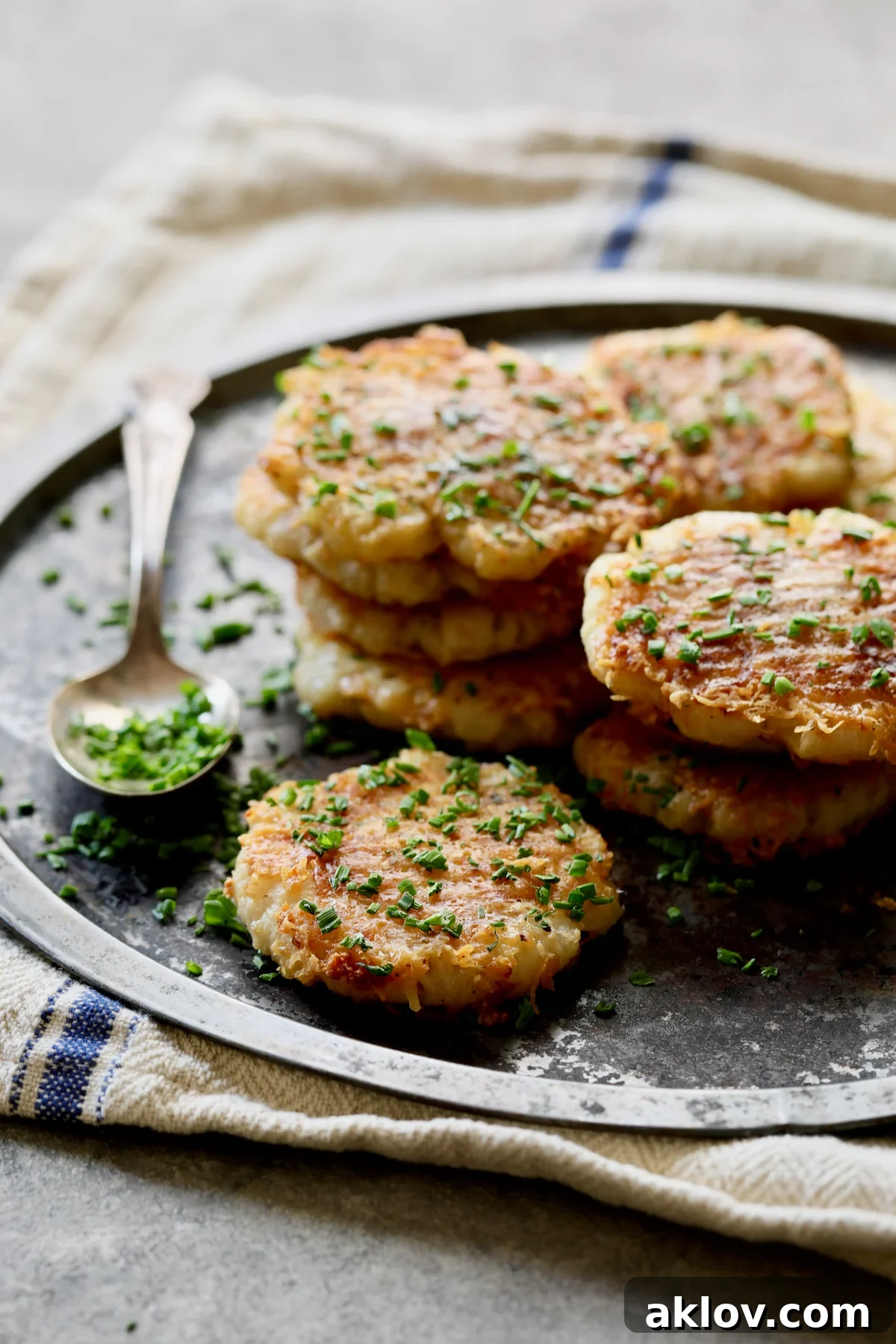 A close-up of stacked Parmesan Crusted Crushed Turnips on a stainless steel tray, generously garnished with freshly chopped chives, ready to be served.