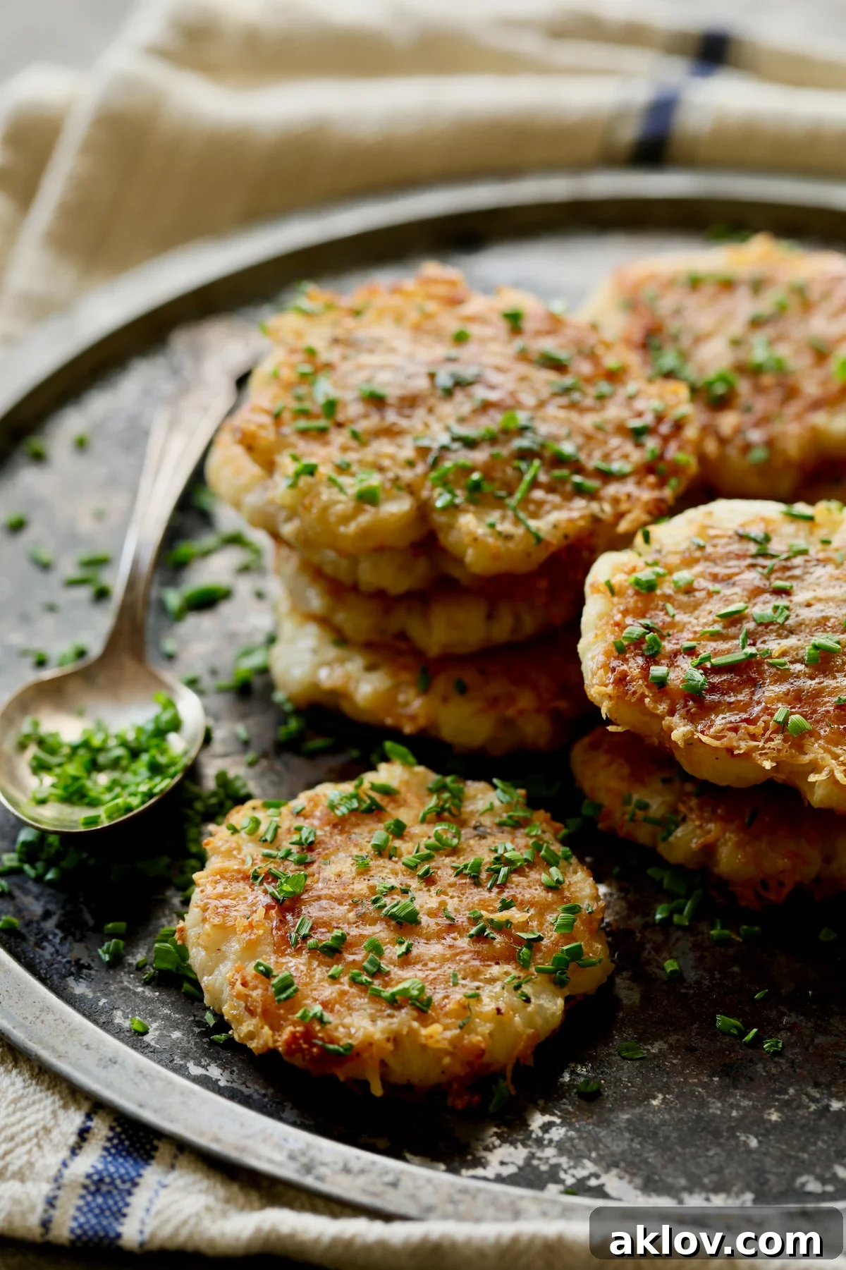A close-up of Parmesan Crusted Crushed Turnips on a stainless steel tray, with a spoonful of fresh chives spilling over, ready to enhance the flavor.