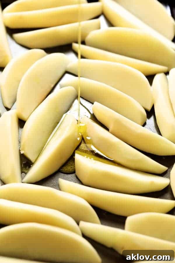 Photo of raw potato wedges on a baking sheet being generously drizzled with olive oil, ready for seasoning and baking.