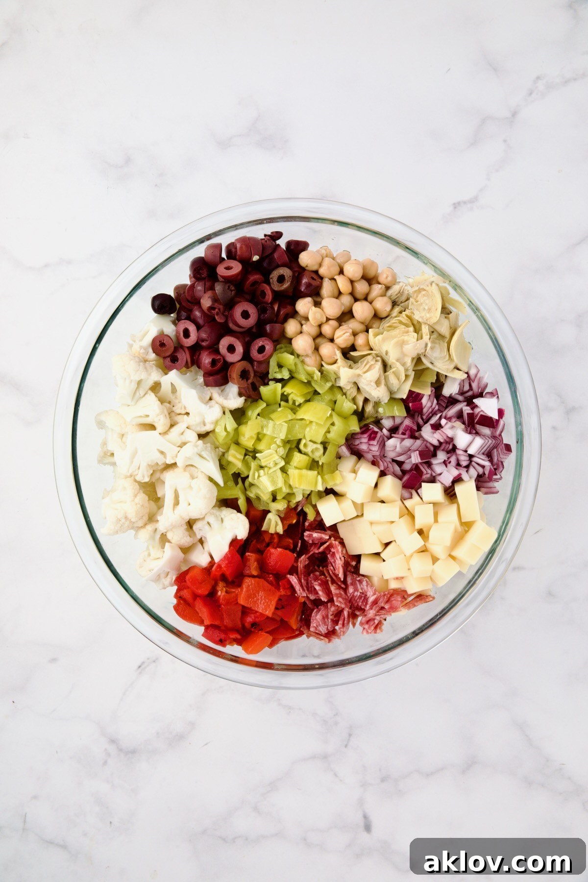All the prepped ingredients for Cauliflower Antipasto Salad, including cauliflower, red onion, roasted red pepper, chickpeas, artichoke hearts, olives, pepperoncini, cheese, salami, and parsley, gathered in a large glass bowl before being mixed.