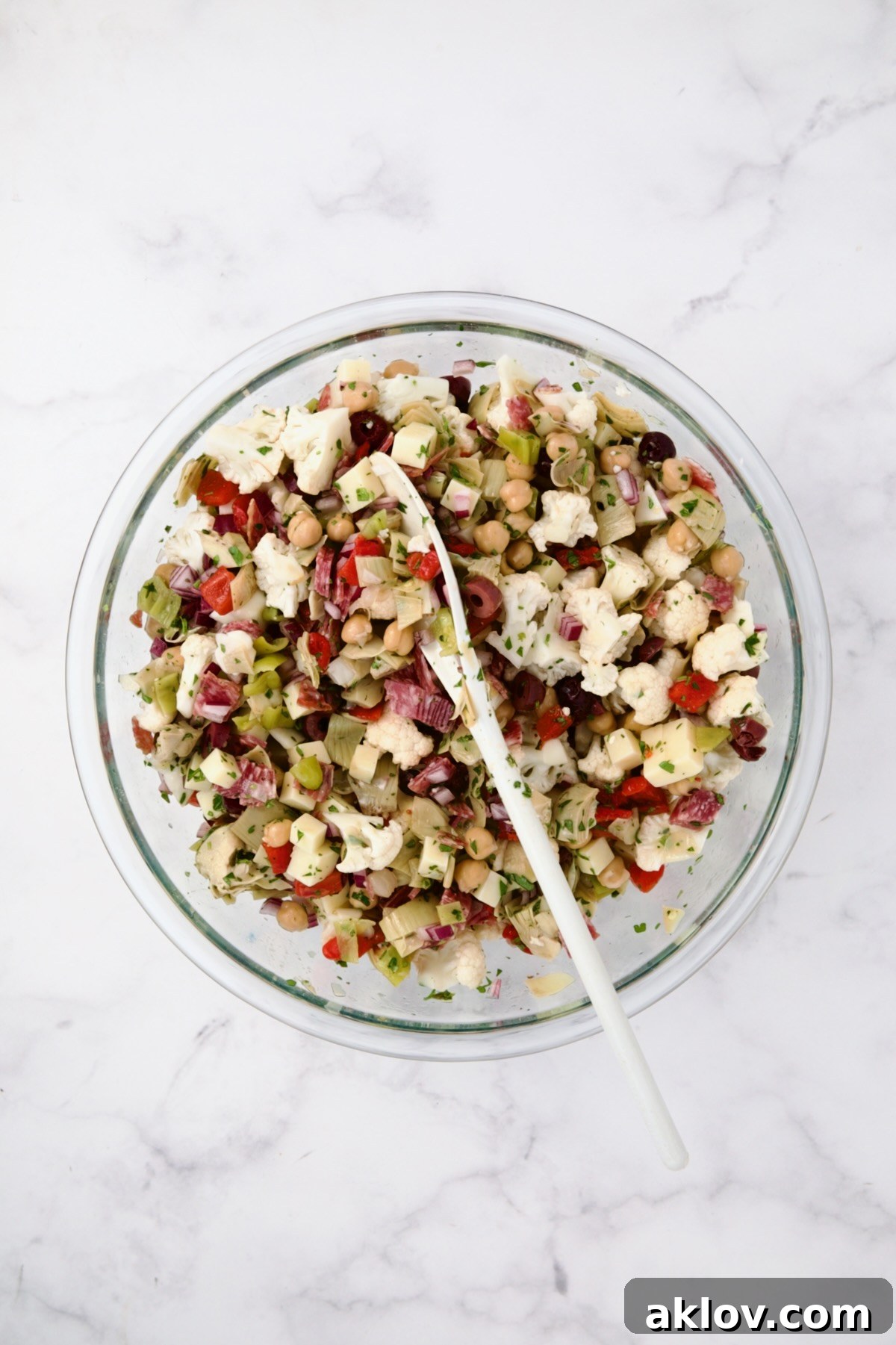The vibrant medley of ingredients for Cauliflower Antipasto Salad, including cauliflower, red onion, roasted red pepper, chickpeas, artichoke hearts, olives, pepperoncini, cheese, salami, and parsley, gently tossed together in a large glass bowl.