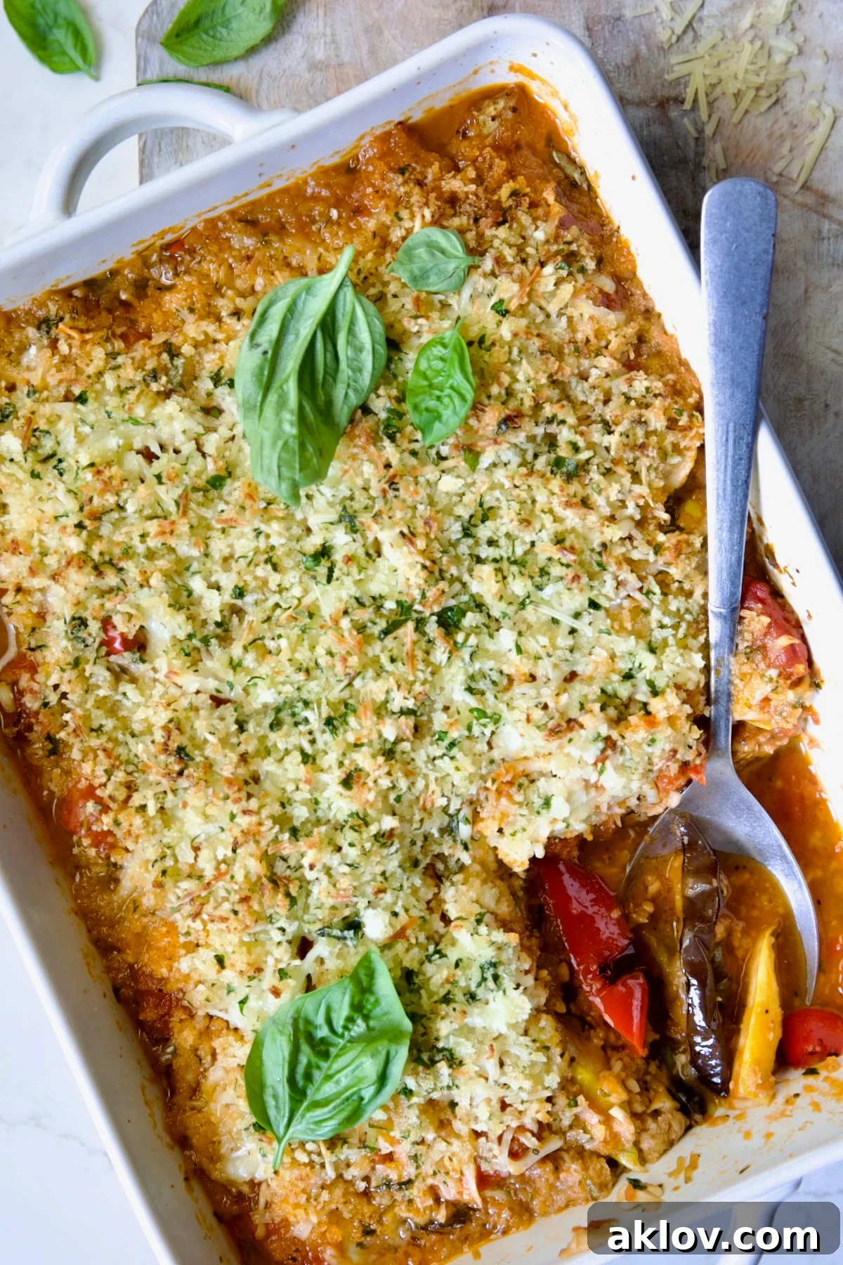 A large white baking dish filled with golden-brown Summer Vegetable Parmigiana, featuring visible layers of grilled vegetables, melted cheese, and marinara sauce, with a serving spoon lifting a generous portion.