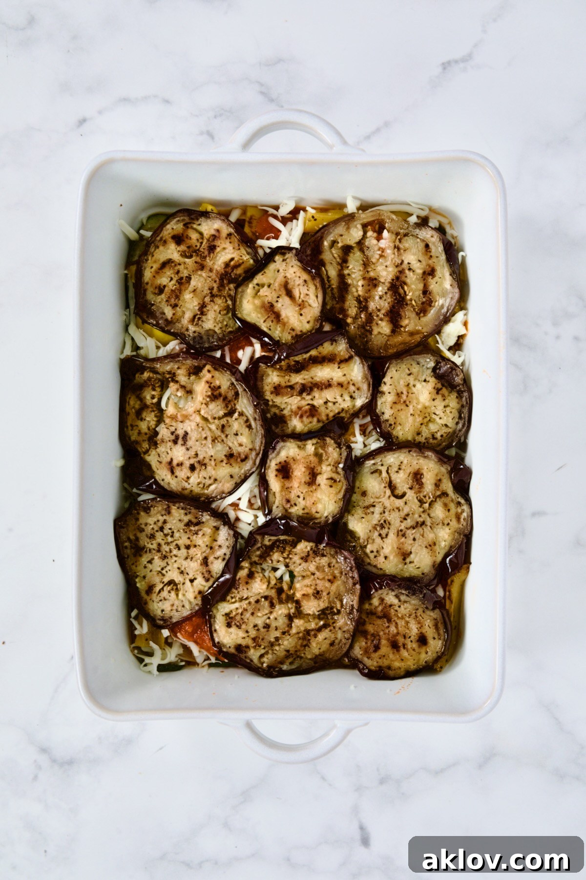 A savory layer of grilled eggplant slices meticulously arranged over sauce and cheese in a white rectangular baking dish, ready for the next delicious additions to the parmigiana.
