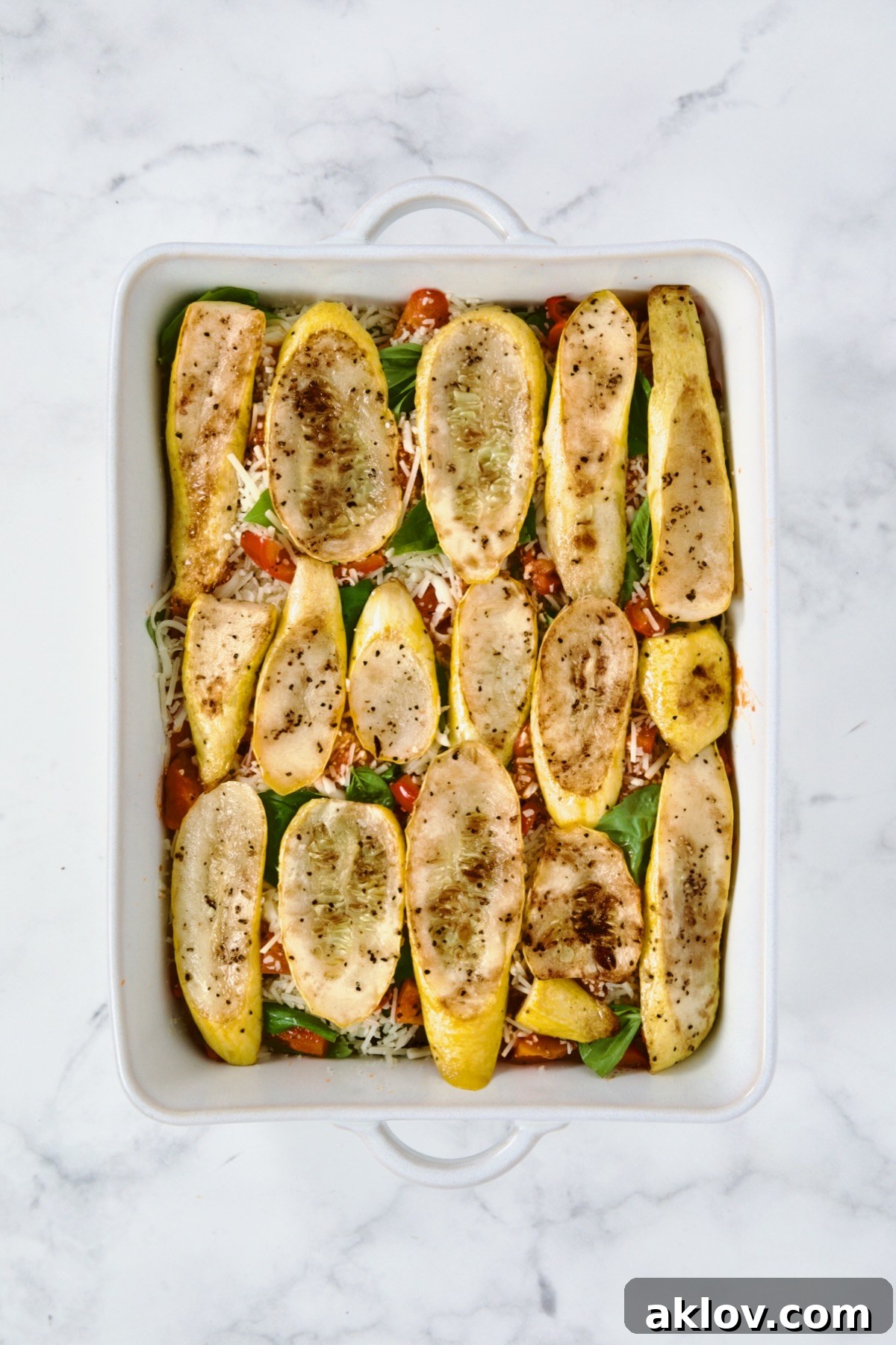 The final vegetable layer of grilled yellow squash slices arranged over sauce and cheese in a white rectangular baking dish, completing the vibrant stack for the Summer Vegetable Parmigiana.