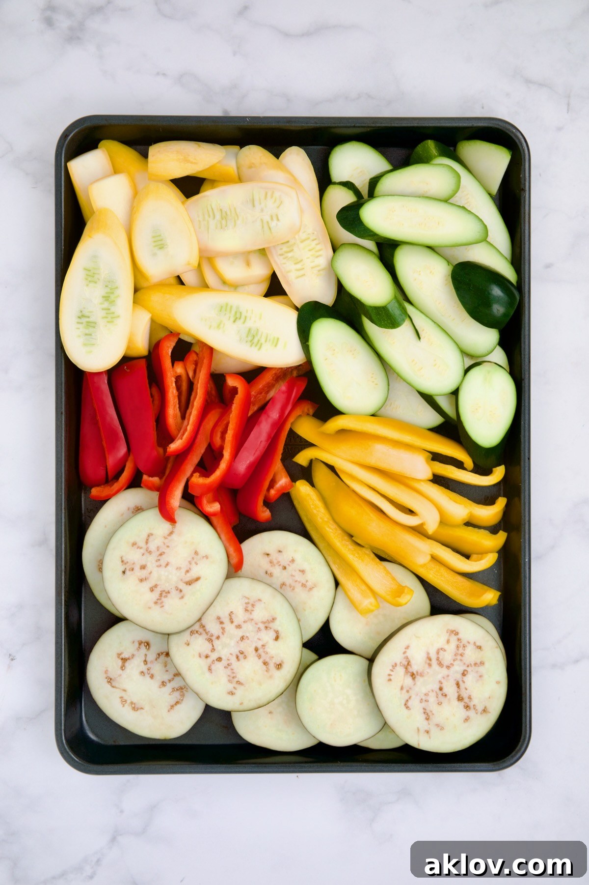 Raw, thinly sliced yellow squash, zucchini, red bell pepper, yellow bell pepper, and eggplant uniformly arranged on a rimmed sheet pan, ready for grilling or roasting.