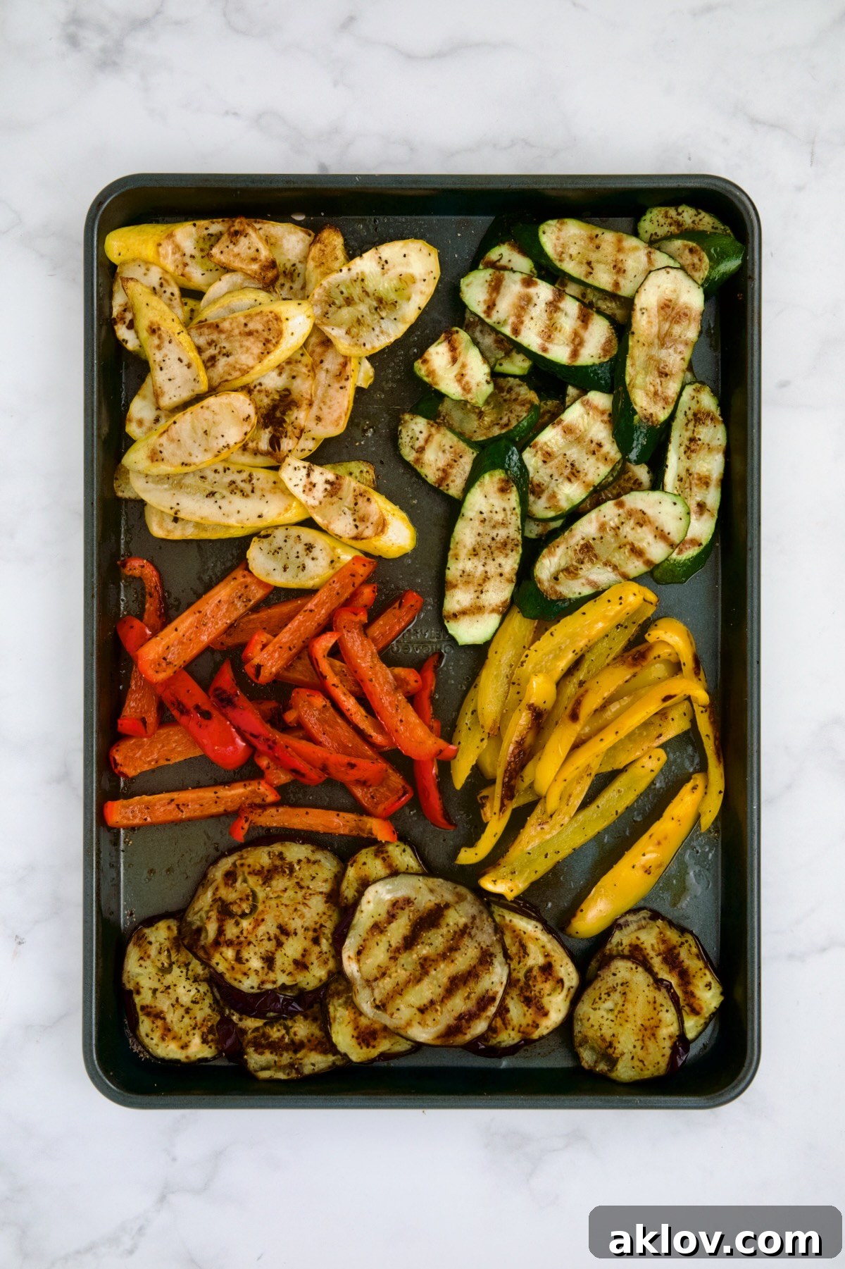 Beautifully grilled yellow squash, zucchini, red bell pepper, yellow bell pepper, and eggplant, showing distinct char marks, cooled on a rimmed sheet pan after cooking.