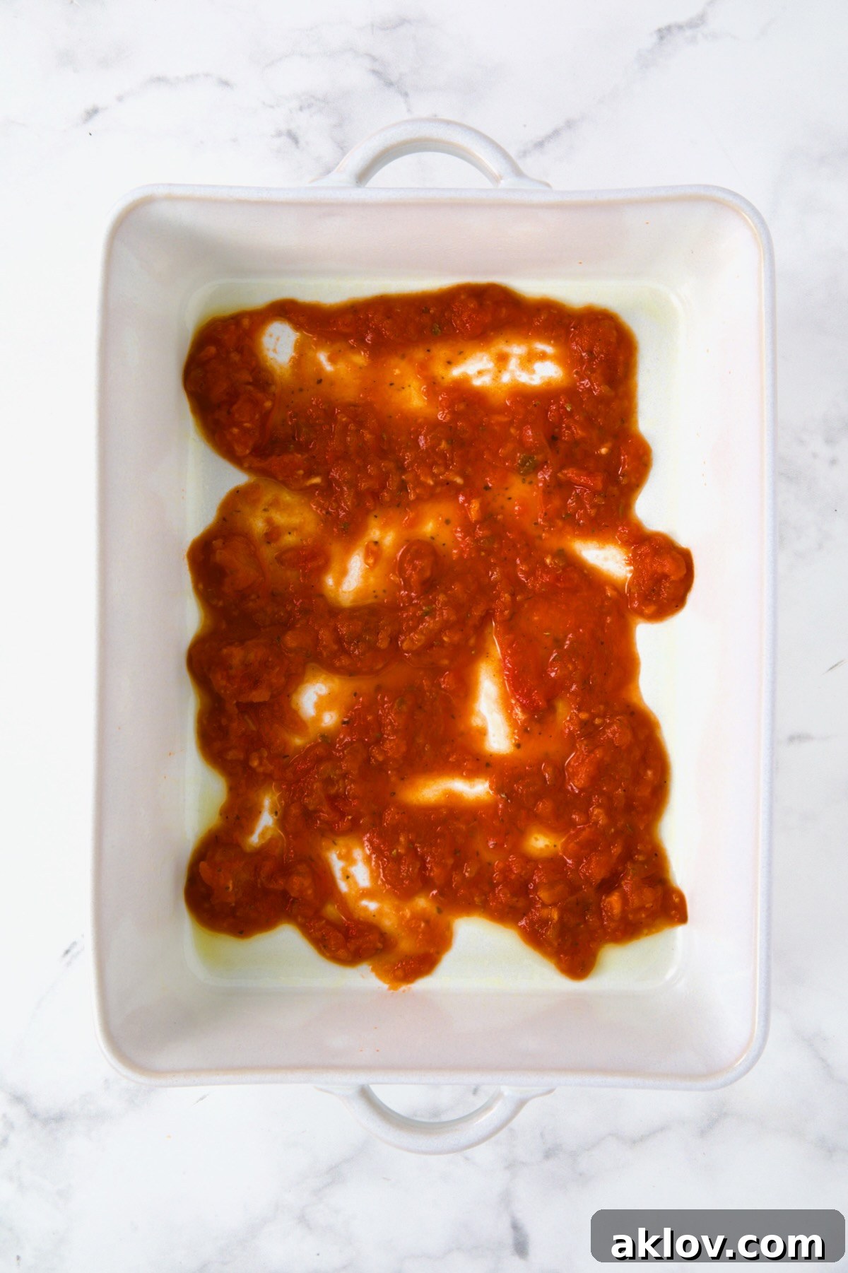 A vibrant red layer of marinara sauce spread across the bottom of a white rectangular baking dish, forming the base for the Summer Vegetable Parmigiana.