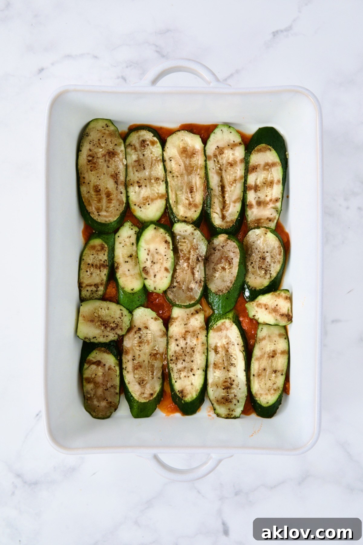 A neatly arranged layer of grilled zucchini slices covering the marinara sauce base in a white rectangular baking dish, marking the first vegetable layer of the Parmigiana.