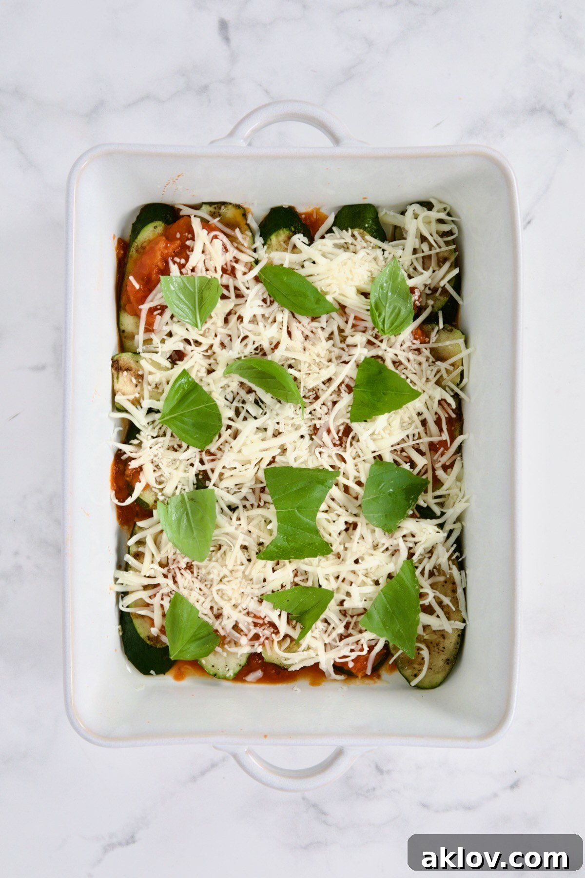 A tantalizing layer of marinara sauce, melted mozzarella and Parmesan cheeses, and fresh green basil leaves artfully spread over grilled vegetables in a white rectangular baking dish.