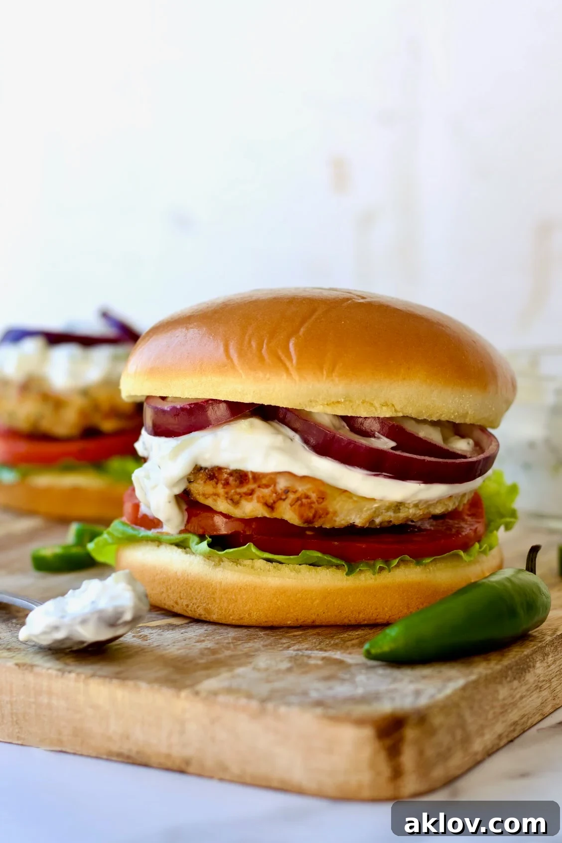 Flavorful Ground Chicken Burgers with Spicy Tzatziki sauce on a rustic wooden cutting board, with a spoon of tzatziki and a fresh jalapeno beside a burger.