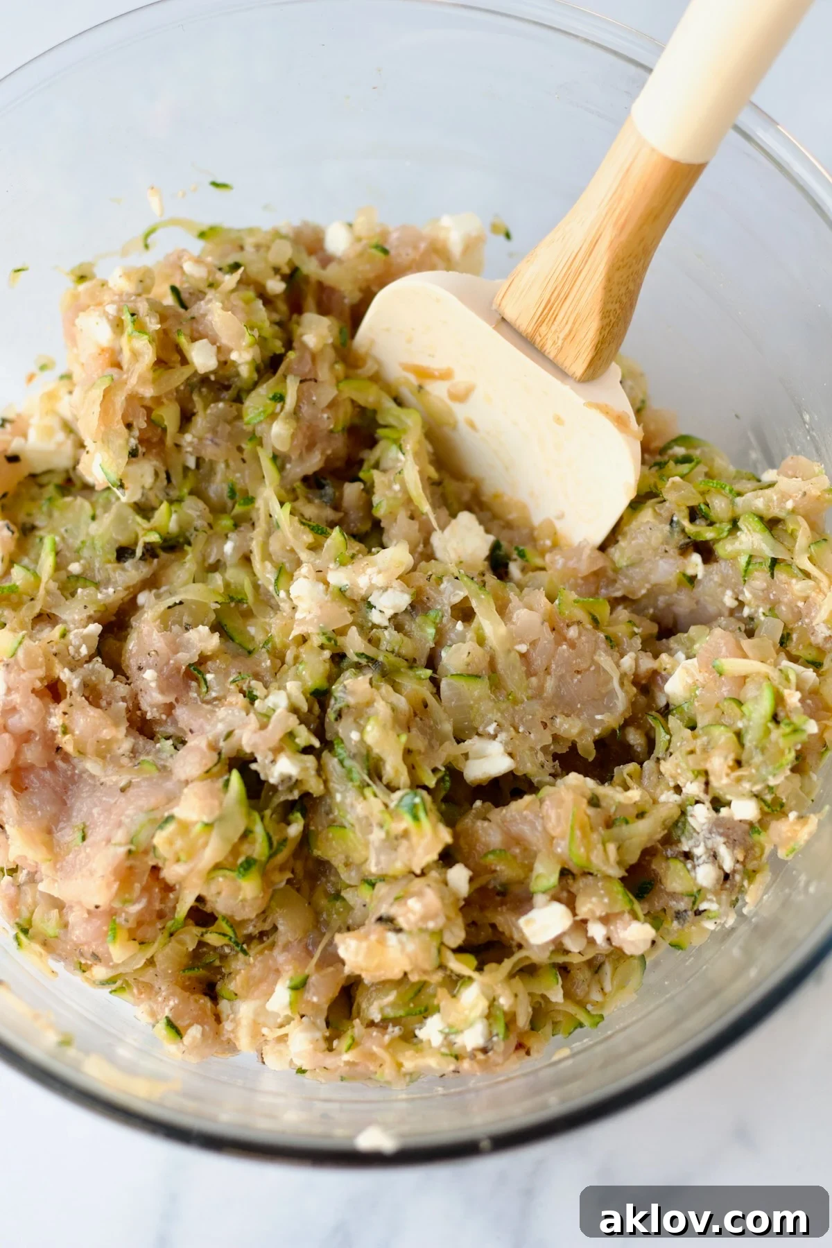 The ground chicken burger ingredients are gently combined in a glass bowl using a rubber spatula, ensuring even distribution without overmixing.
