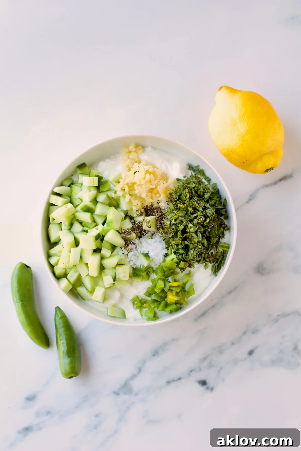 Bowls of ingredients for the spicy tzatziki sauce, including shredded cucumber, diced jalapeno, and fresh mint, are arranged on a kitchen countertop.