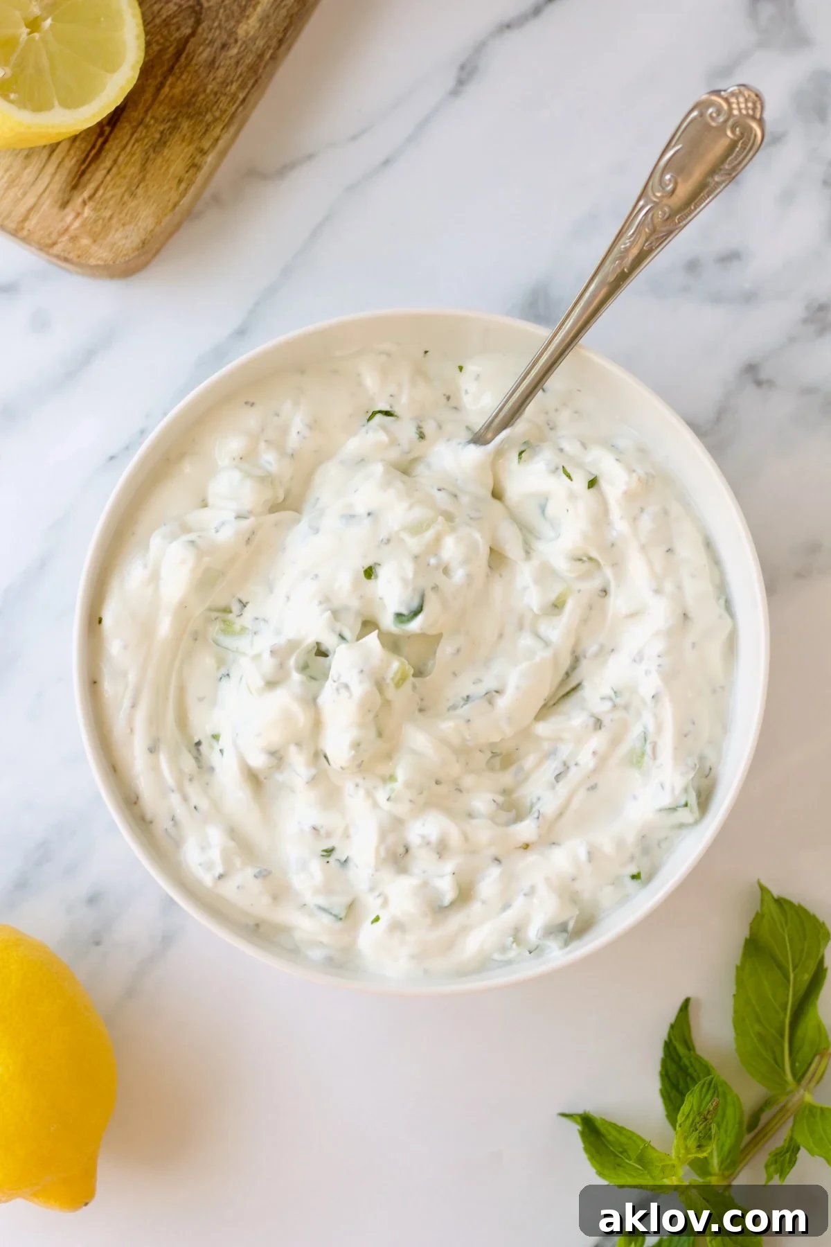 A serving of creamy spicy tzatziki sauce rests in a pristine white bowl with an ornate spoon on a stylish marble countertop.