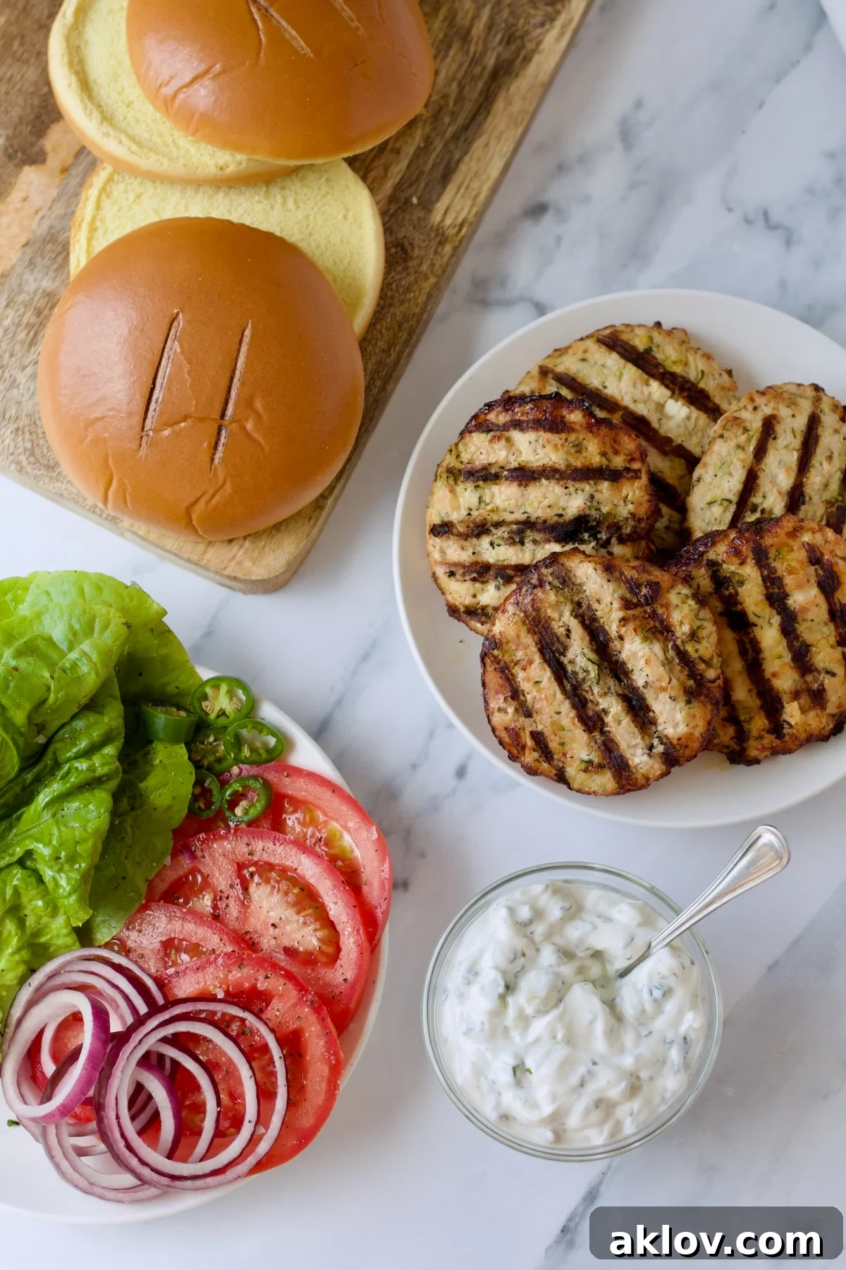All the components for assembling Ground Chicken Burgers with Spicy Tzatziki, including grilled patties, fresh vegetables, buns, and sauce, are laid out and ready.