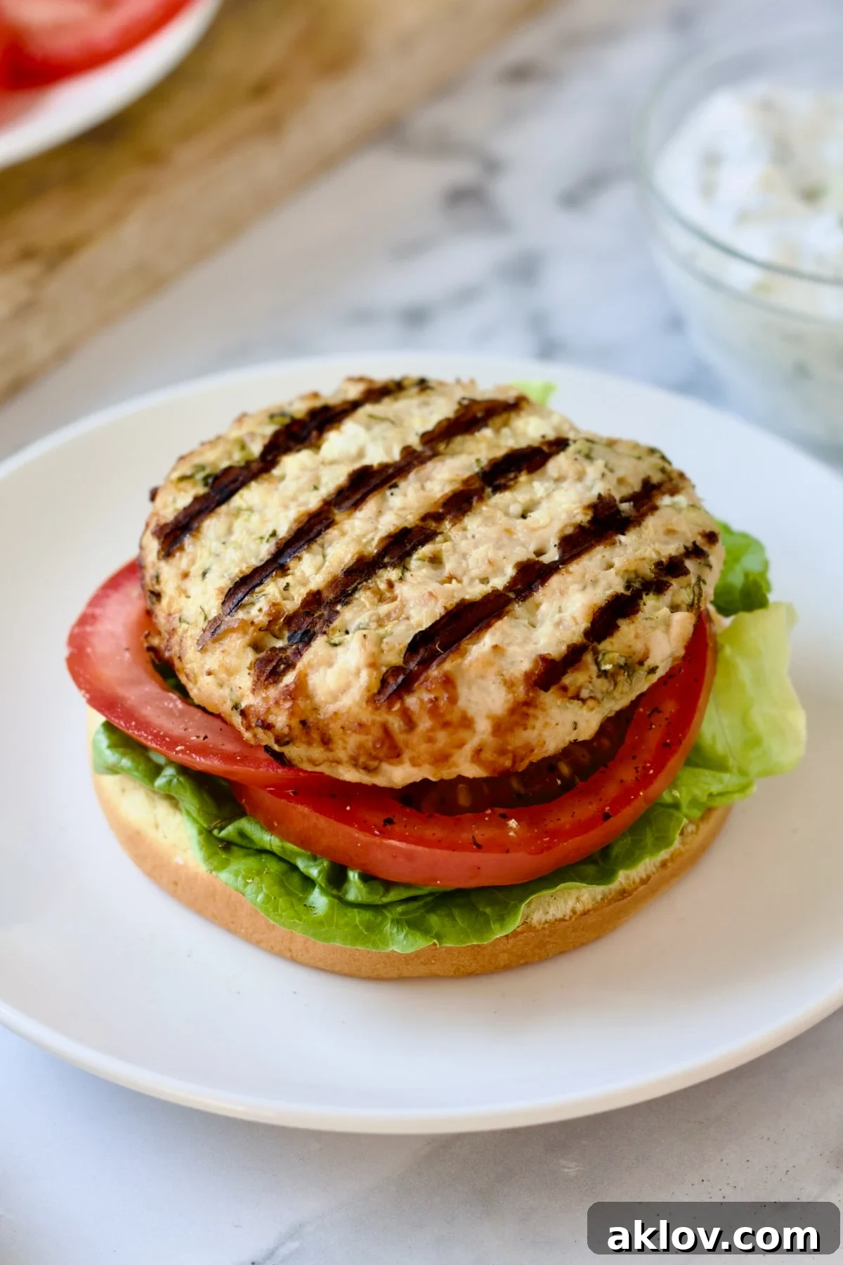 A freshly grilled ground chicken burger, topped with crisp lettuce and a ripe tomato slice, awaits its spicy tzatziki and red onion on a brioche bun.