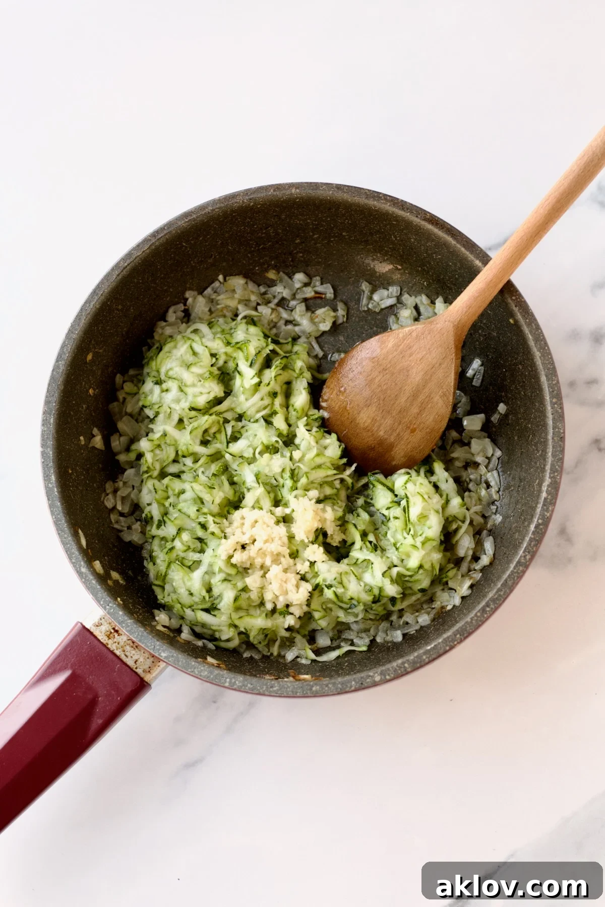 Shredded zucchini and minced garlic are mixed into the sautéed onions in a nonstick skillet, stirred with a wooden spoon to combine flavors.