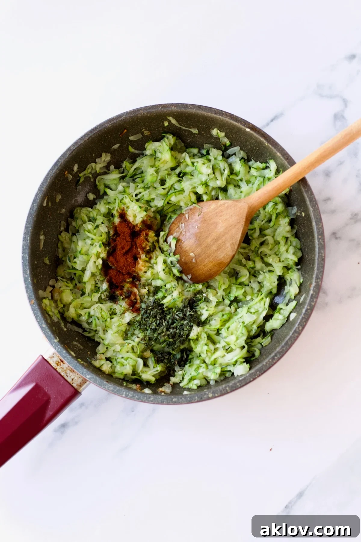 Cayenne pepper and fresh oregano are being stirred into the sautéed zucchini and onion mixture in a nonstick skillet with a wooden spoon.