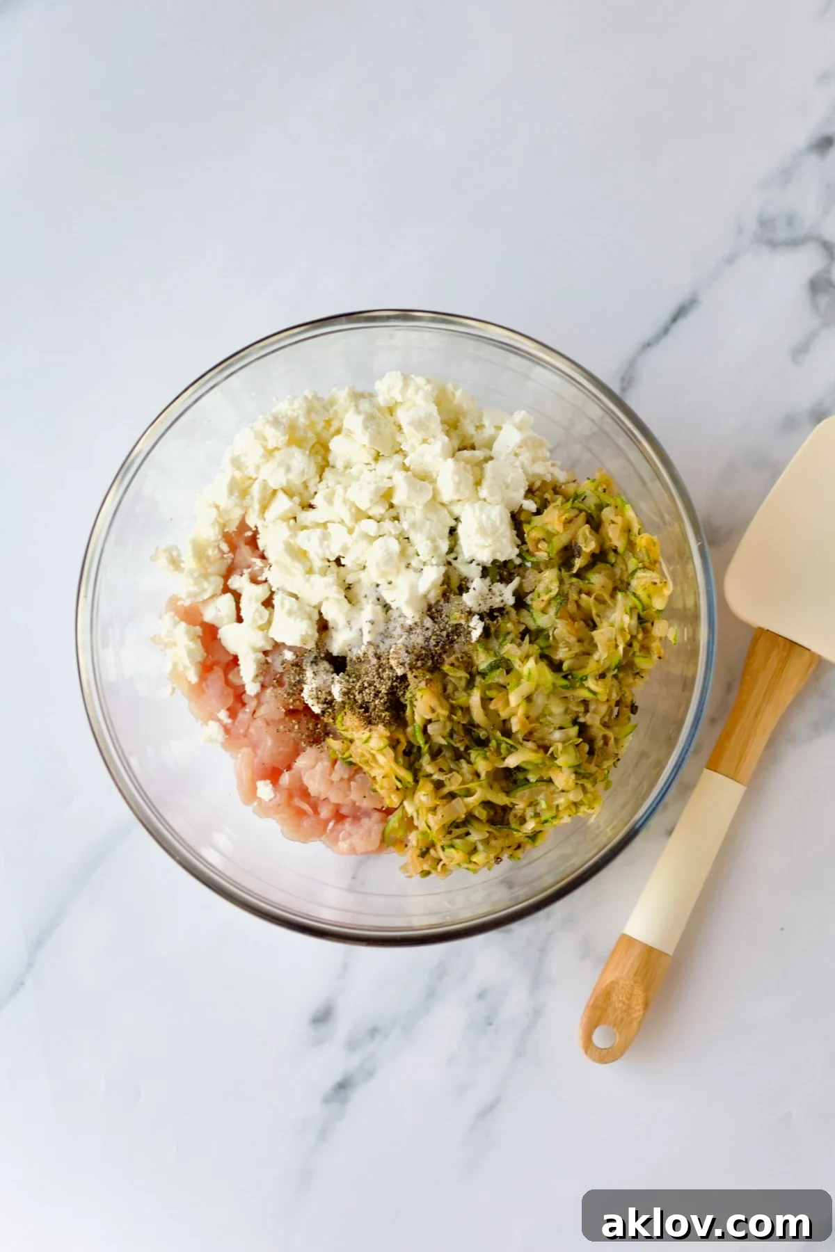 All the essential ingredients for the chicken burgers, including ground chicken, feta, and the cooled vegetable mixture, are placed in a clear glass bowl, ready for mixing.