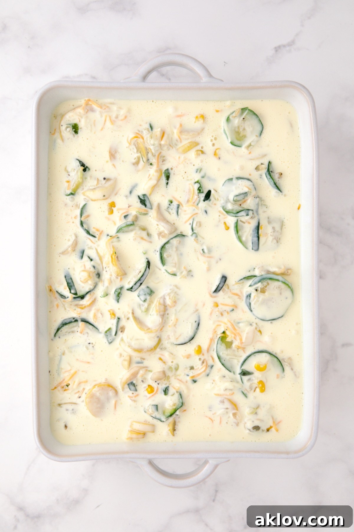 The creamy custard mixture being poured over the vegetable and cheese blend in a white rectangular baking dish.