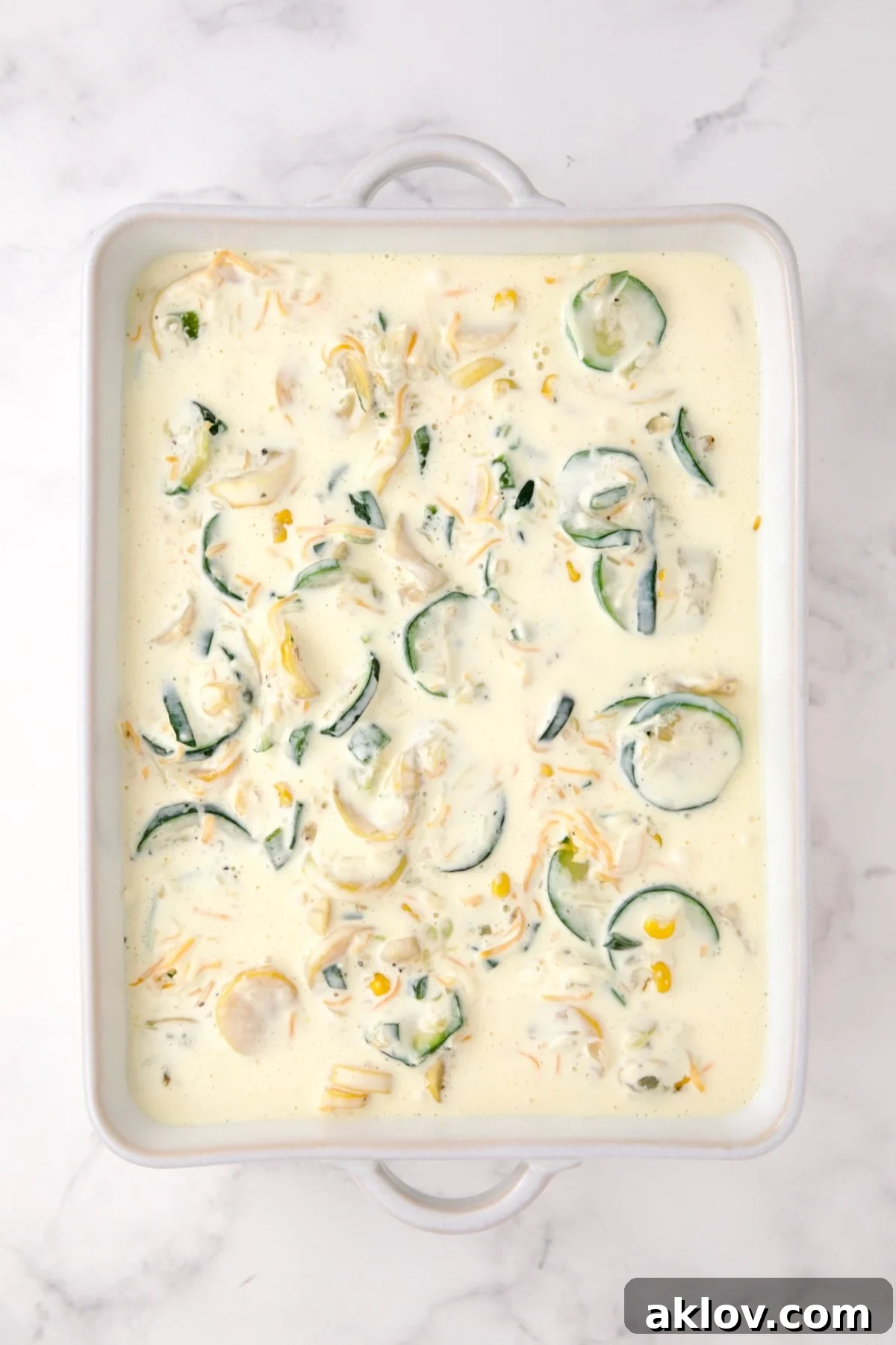 Cooked zucchini, yellow squash, poblano, and corn in a white rectangular baking dish with shredded cheese and the custard mixture added.