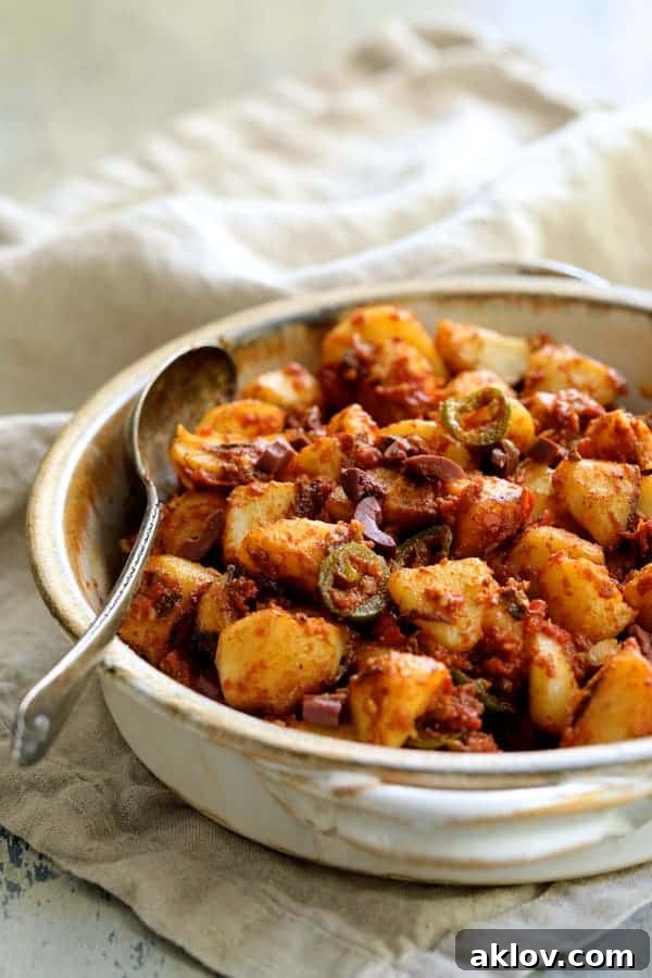 A close-up shot of Patatas Bravas Chilenos elegantly arranged in a rustic clay serving dish, nestled on a textured beige towel. The golden-brown potatoes are generously coated in a vibrant, rich red sauce, hinting at their spicy and tangy flavors, ready to be enjoyed.
