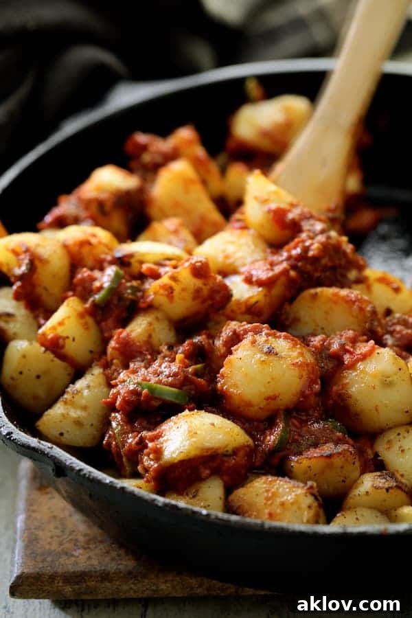 A close-up shot captures the moment golden-brown potatoes are meticulously stirred with a rich, red sauce in a cast-iron skillet, a wooden spoon gently folding the ingredients together. The vibrant sauce coats each potato, highlighting the textural contrast and enticing aromas.