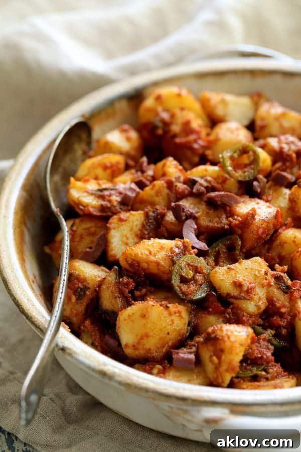 A close-up captures a clay bowl brimming with Patatas Bravas Chilenos, each golden potato cube generously coated in a rich, spicy tomato sauce. A serving spoon rests invitingly in the dish, hinting at the deliciousness within, adorned with a scattering of vibrant garnishes.