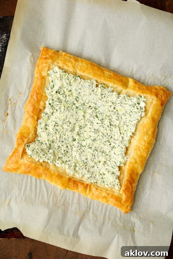 Photo of a baked puff pastry crust on parchment paper with the creamy herbed ricotta mixture evenly spread over its center.