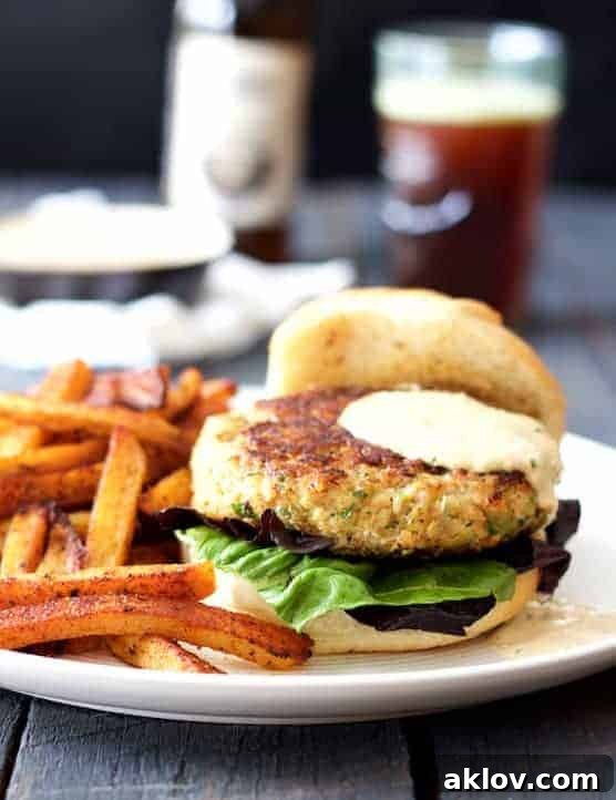 Close-up photo of a beautifully assembled Shrimp Burger with Jalapeño Remoulade, served alongside golden sweet potato fries on a white plate, with a glass of beer subtly blurred in the background. The burger features a thick, golden-brown shrimp patty, fresh lettuce, and a generous dollop of creamy remoulade.