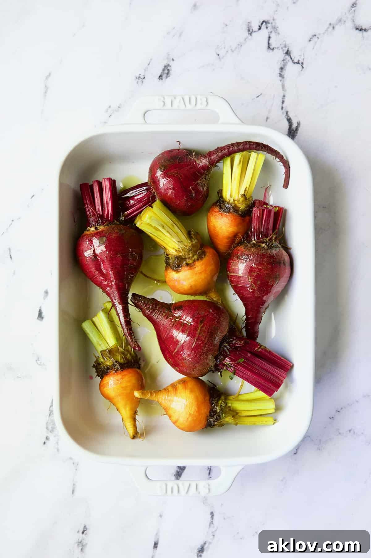Uncooked red and yellow beets in white baking dish drizzled with oil.