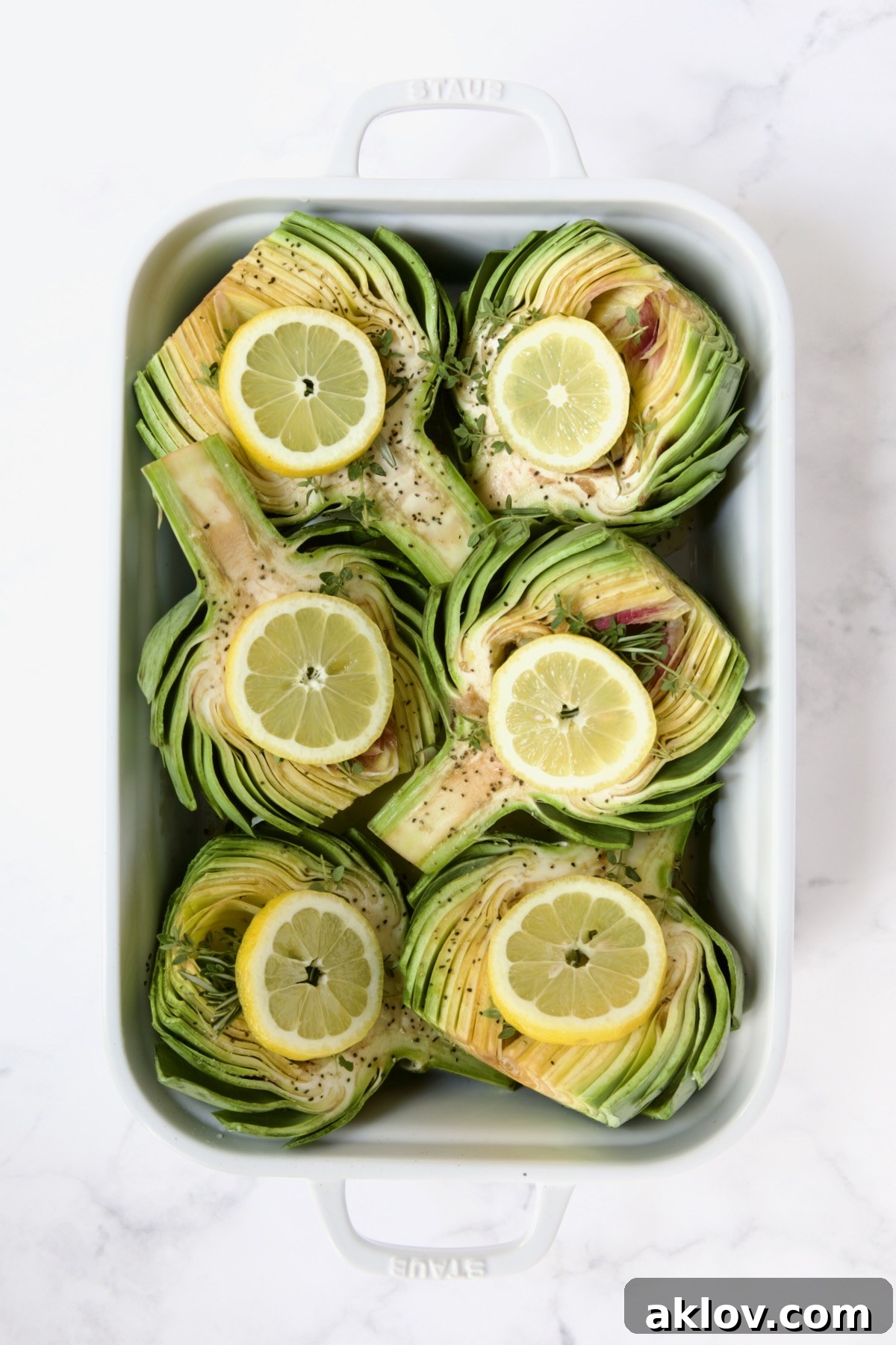 Buttery Garlic Roasted Artichokes 11 Six artichoke halves in a white roasting pan with lemon slices set atop the herbs and garlic cloves.