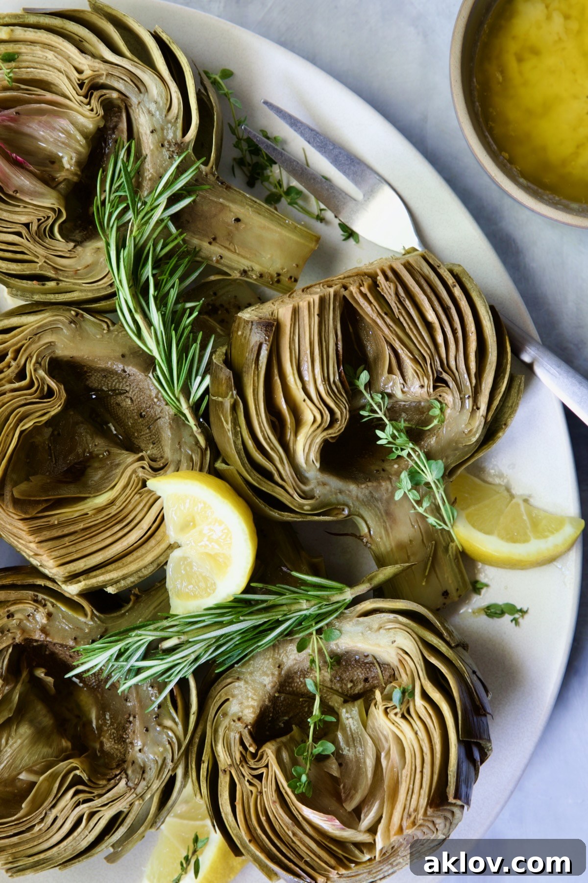 Buttery Garlic Roasted Artichokes 21 Roasted Artichokes with Garlic Butter on white oval platter garnished with herb sprigs and lemon wedges.