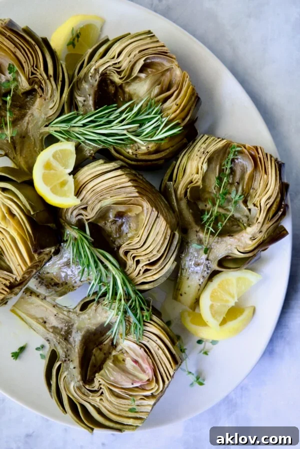 Buttery Garlic Roasted Artichokes 22 Roasted Artichokes with Garlic Butter on white oval platter garnished with lemon wedges and herb sprigs.