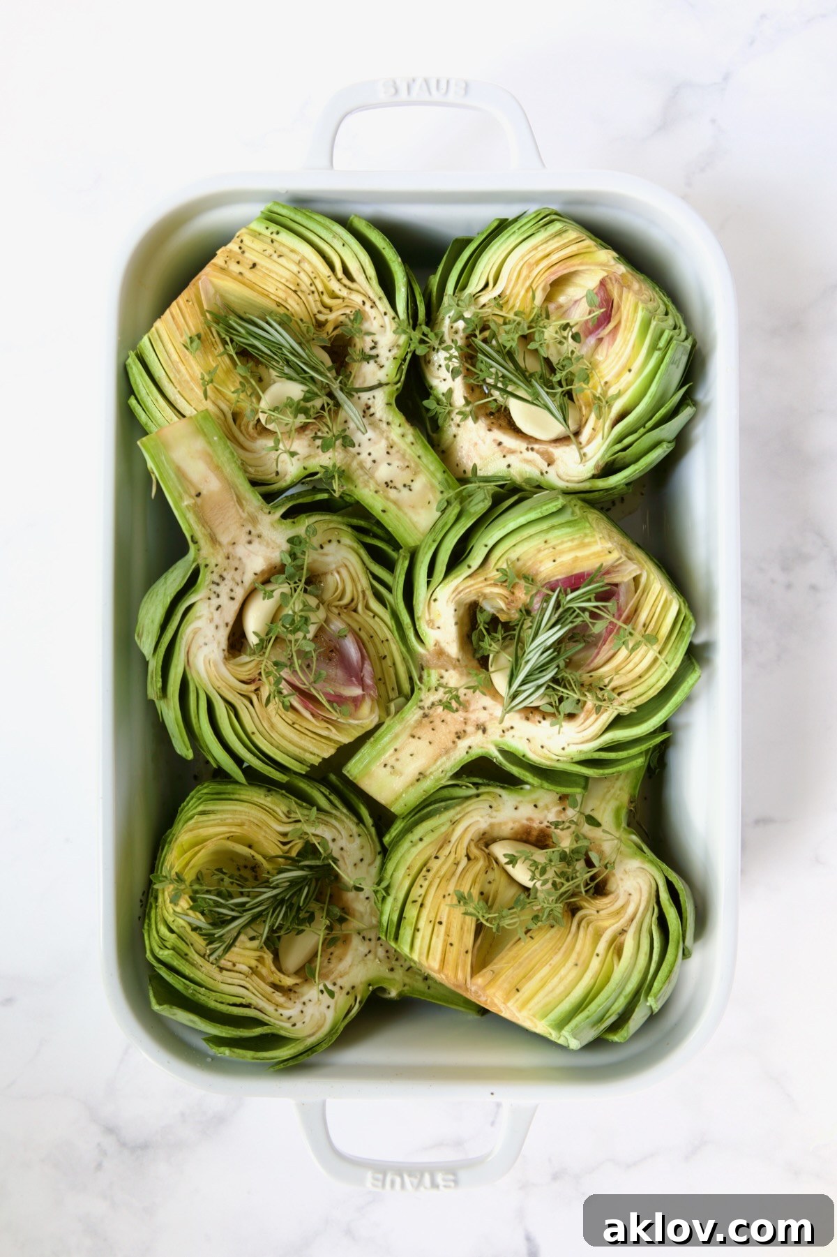 Buttery Garlic Roasted Artichokes 10 Six artichoke halves in a white roasting pan with garlic and herbs on top of them.