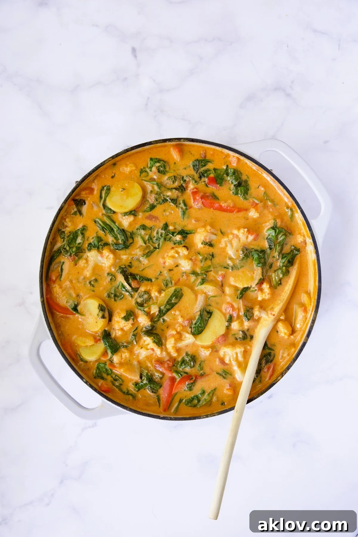 Finished vegetable curry in a white enamel cast iron skillet, being stirred with a white spoon, showing its creamy texture.