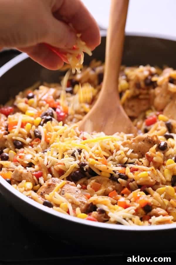 A hand sprinkles shredded Mexican blend cheese over the hot Southwestern Chicken Orzo and Black Bean Skillet in the pan, preparing it for its final melty touch.