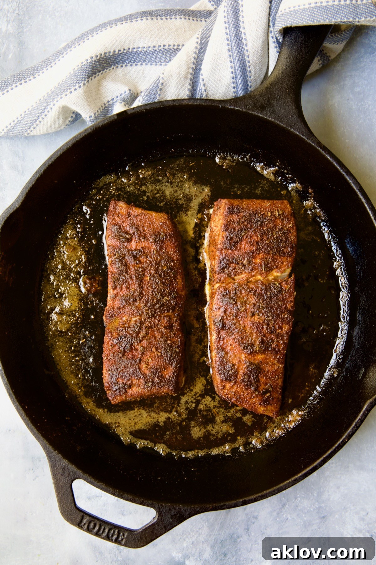 Two pieces of halibut coated with blackening seasoning in cast iron skillet after being cooked with blue striped towel wrapped around the handle.